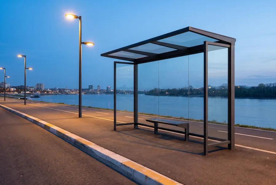 Generated scene: an empty glass bus shelter with a bench beside a riverside road at blue hour, street lights on and a city across the river.